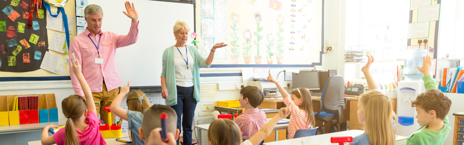Two teachers in a classroom standing in front of their class with their hands raised
