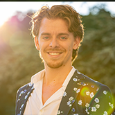 Smiling man in a floral-patterned blazer and open-collar shirt outdoors, lit by warm sunlight with lens flare.