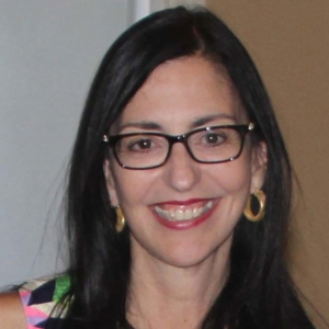 Headshot of a smiling woman with long dark hair, black rectangular glasses, and gold hoop earrings.