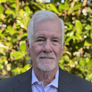 Head-and-shoulders portrait of an older man with white hair and a trimmed beard, wearing a suit jacket and light shirt, outdoors with leafy greenery in the background.
