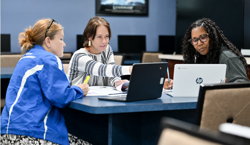 Three people sit around a table in a classroom or office, collaborating over open laptops and papers while one points at a screen.