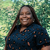Smiling person with long braids wearing a dark patterned top, standing outdoors in front of leafy greenery.