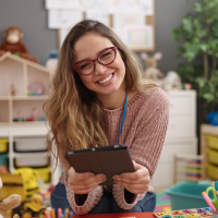 A young teacher smiles while sitting in a preschool classroom.