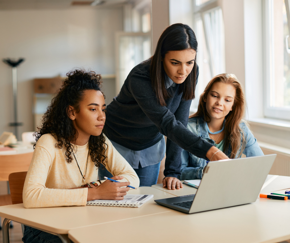 Teacher stands between two students at a desk to help them with their work.