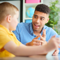 A teacher sits at a desk and talks to a young student.