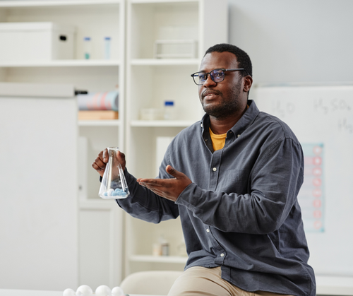 A high school chemistry teacher lectures at the front of a classroom while holding a beaker.