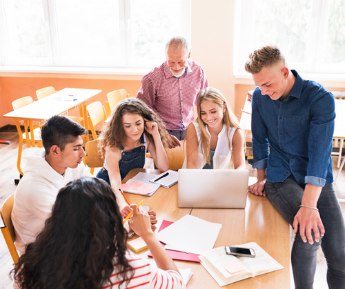 A teacher stands at a table with a group of students working on a project.