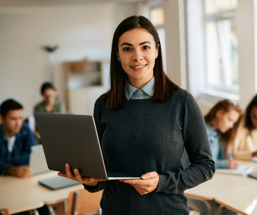 A Social Studies teacher stands in her classroom holding her laptop while students work at their desks.