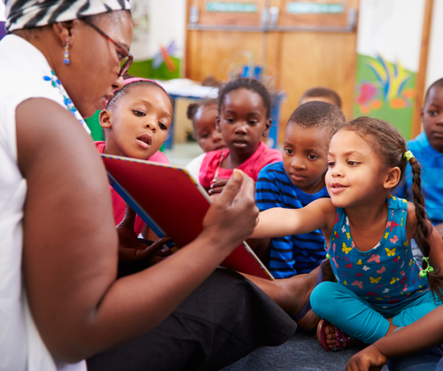 A teacher sits with a group of children reading a book, the children are pointing at the pages.