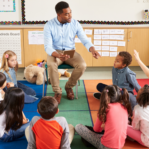 A male teacher sitting on a chair in a classroom surrounded by students sitting on the ground