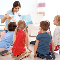 A teacher sits while reading a book to a group of young students.