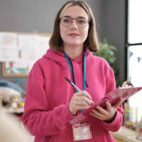 A teacher stands in a classroom holding a clipboard.
