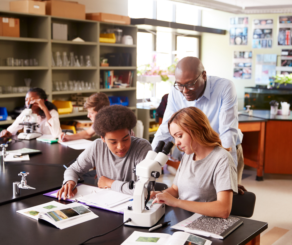 A high school Biology teacher helps students use microscope in a classroom.