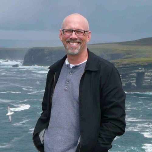 Dr. John White stands on a grassy cliff with water behind him smiling. 
