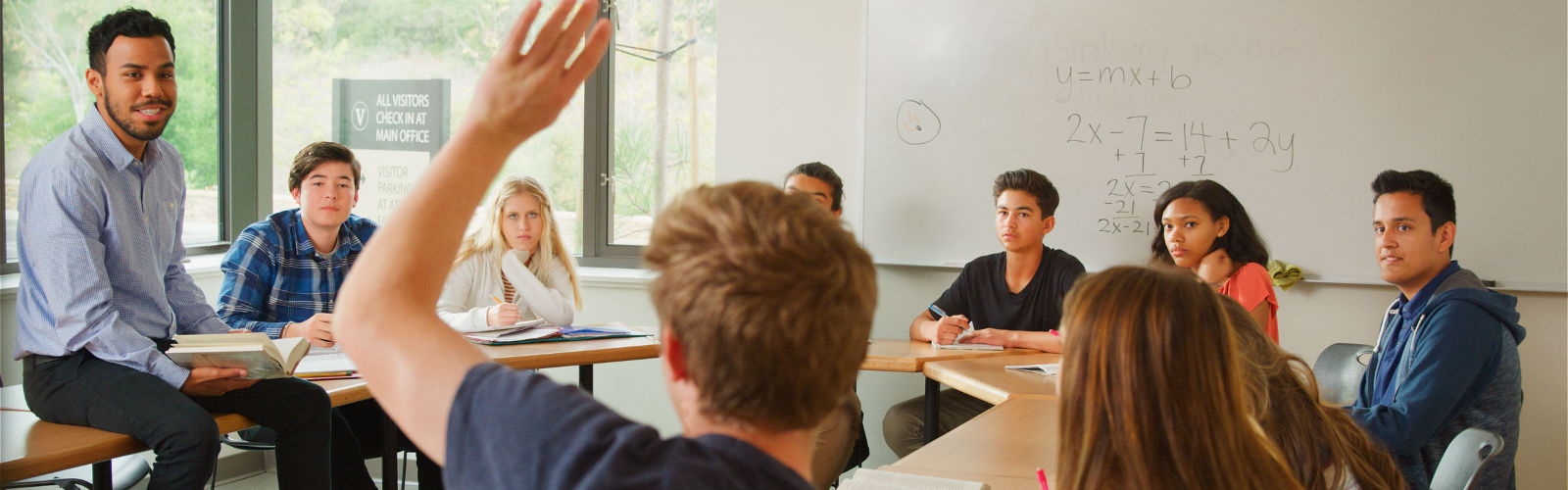 A teacher sits in the middle of a high school math classroom calling on students with their hands raised. 