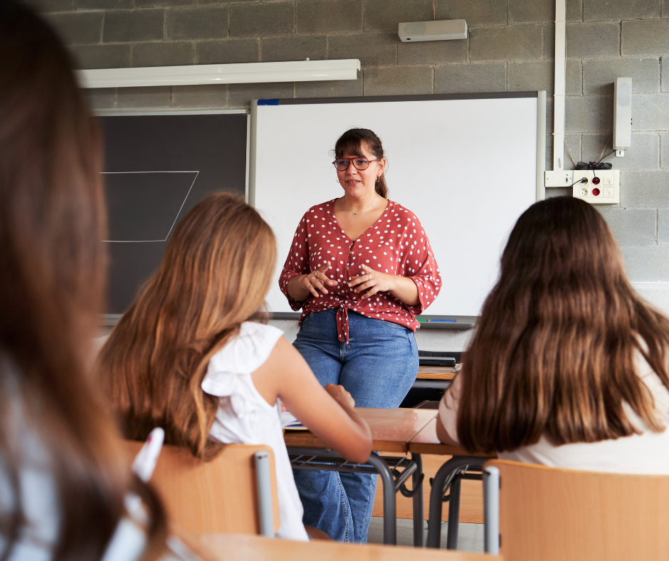 A teacher stands at the front of a high school classroom while students sit in desks.