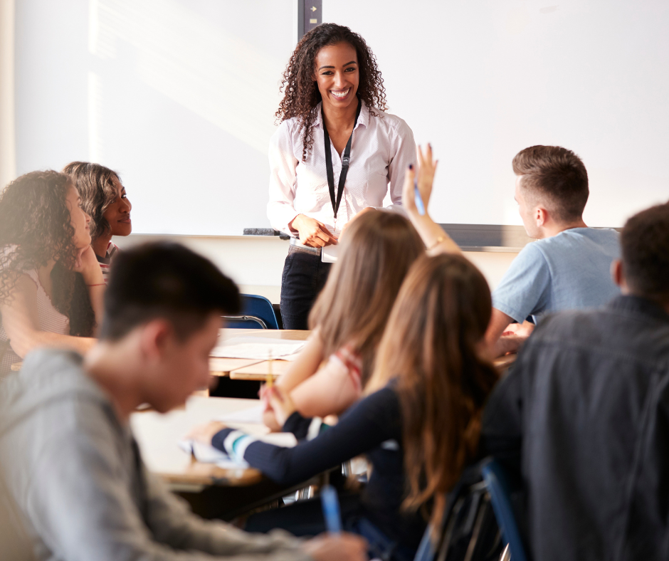 A teacher stands, smiling surrounded by students sitting in a high school social studies classroom.