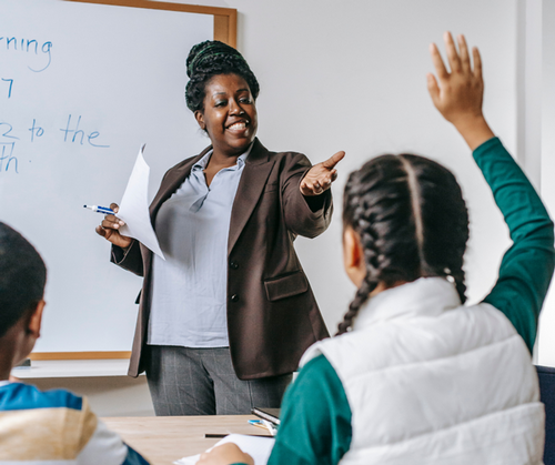 A teacher stands at the front of a classroom calling on students who raised their hands.