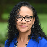 Head-and-shoulders portrait of a woman with curly dark hair and glasses, wearing a blue blazer and smiling against a blurred green outdoor background.