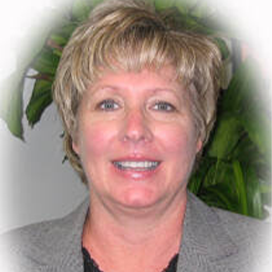Smiling person with short blond hair wearing a gray blazer, with green plant leaves in the background.