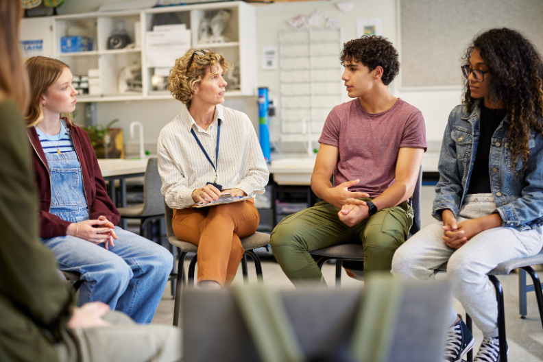 Facilitator with a clipboard leads a small group discussion with teenagers seated in a circle in a classroom.