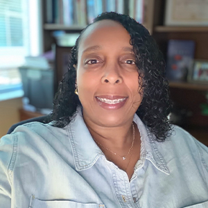 Smiling person with curly hair in a light denim shirt, seated indoors with bookshelves in the background.