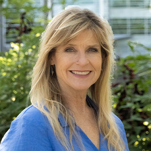 Smiling woman with long blonde hair wearing a blue top, standing outdoors with greenery and a building in the background.