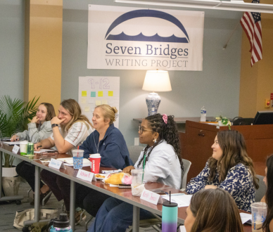 Panelists sit at a long table in a classroom-style room with a “Seven Bridges Writing Project” banner behind them and an American flag to the side.