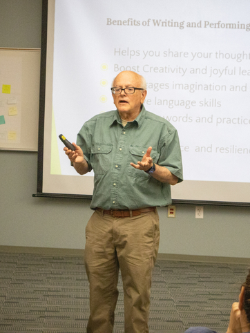 Man giving a presentation in a classroom, holding a remote and gesturing in front of a projected slide.