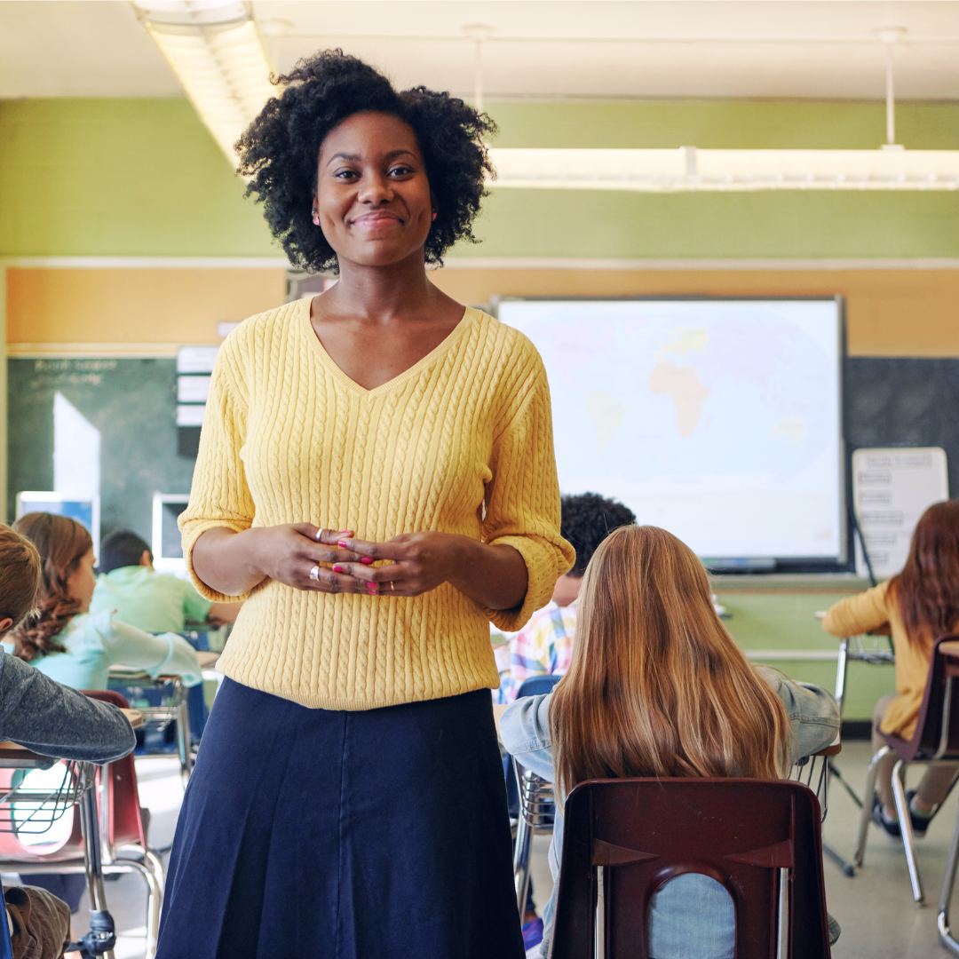 A teacher stands in a classroom smiling. 