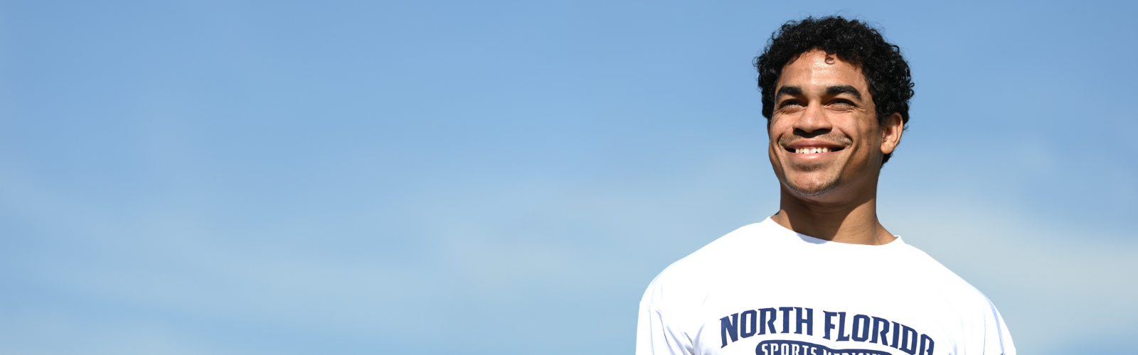 UNF student smiling wearing a white UNF Sports Medicine shirt in front of blue sky