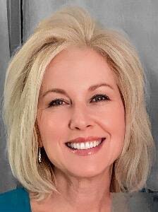 Smiling woman with shoulder-length blonde hair, shown in a close-up portrait against a gray background.