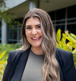 Smiling woman with long wavy hair wearing a dark blazer, standing outdoors near greenery with a modern building in the background.