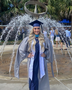 Graduate in cap and gown smiling in front of a fountain with water jets, with palm trees and people in the background.