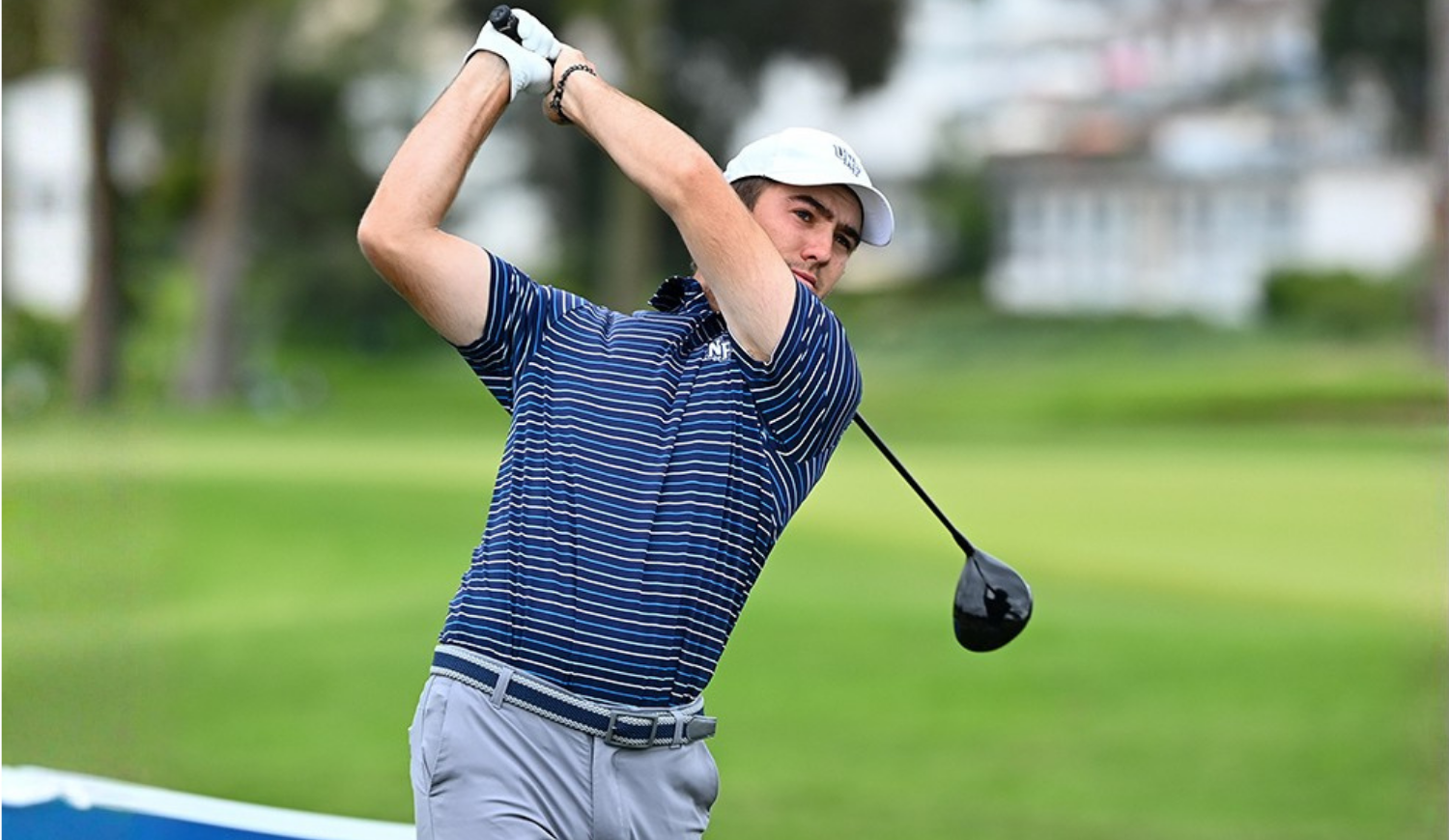 Golfer in a striped shirt and white cap follows through on a tee shot with a driver on a green course.