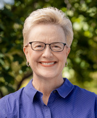 Carolyn Ball in a lavender shirt standing outside in front of trees.
