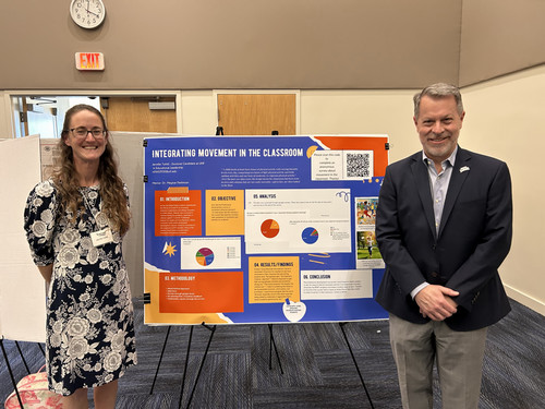 A woman and man pose beside a bright poster on movement in classrooms.