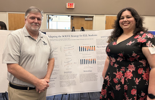 A man and woman stand in front of a poster on ELL student strategies.