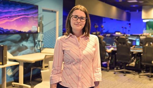 Person wearing glasses and a pink striped shirt stands in a modern office workspace with computer stations in the background.