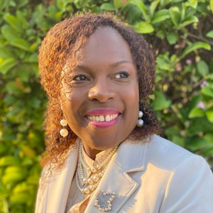 Headshot of Dunia Jean Baptiste smiling outdoors with greenery in the background.