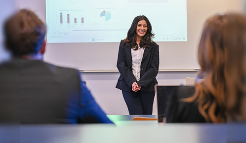 Business presenter smiling at the front of a meeting room, with a projected slide showing charts in the background and attendees in the foreground.