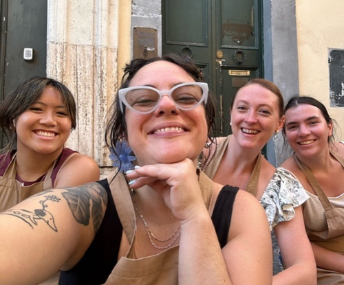 Four women in aprons smile together outside a building entrance