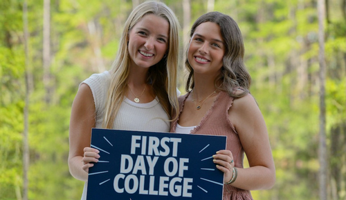 Two young women smiling outdoors and holding a sign that reads “First Day of College.”