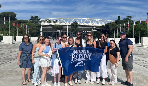 A group of students and faculty stand holding a UNF banner in Italy.