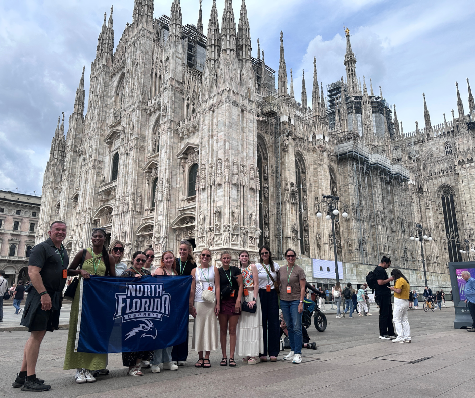 A group of students and faculty stand holding a UNF banner in Italy.