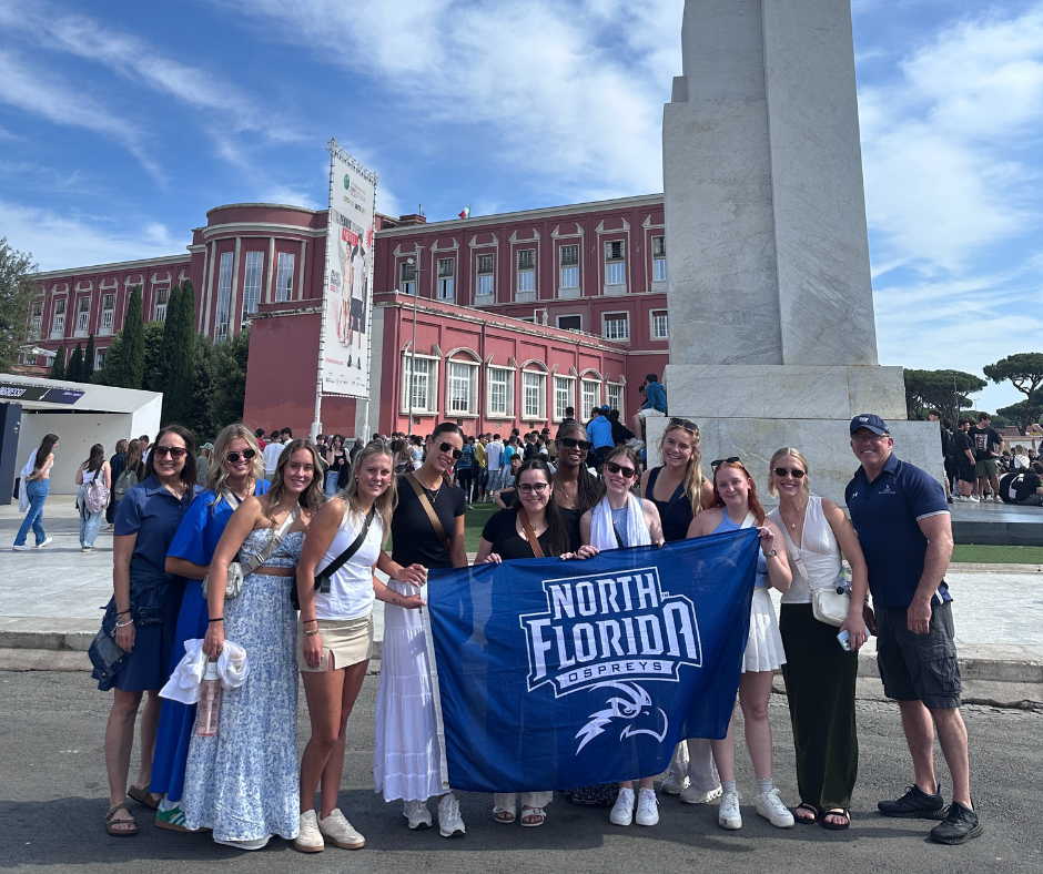 A group of students and faculty stand holding a UNF banner in Italy.