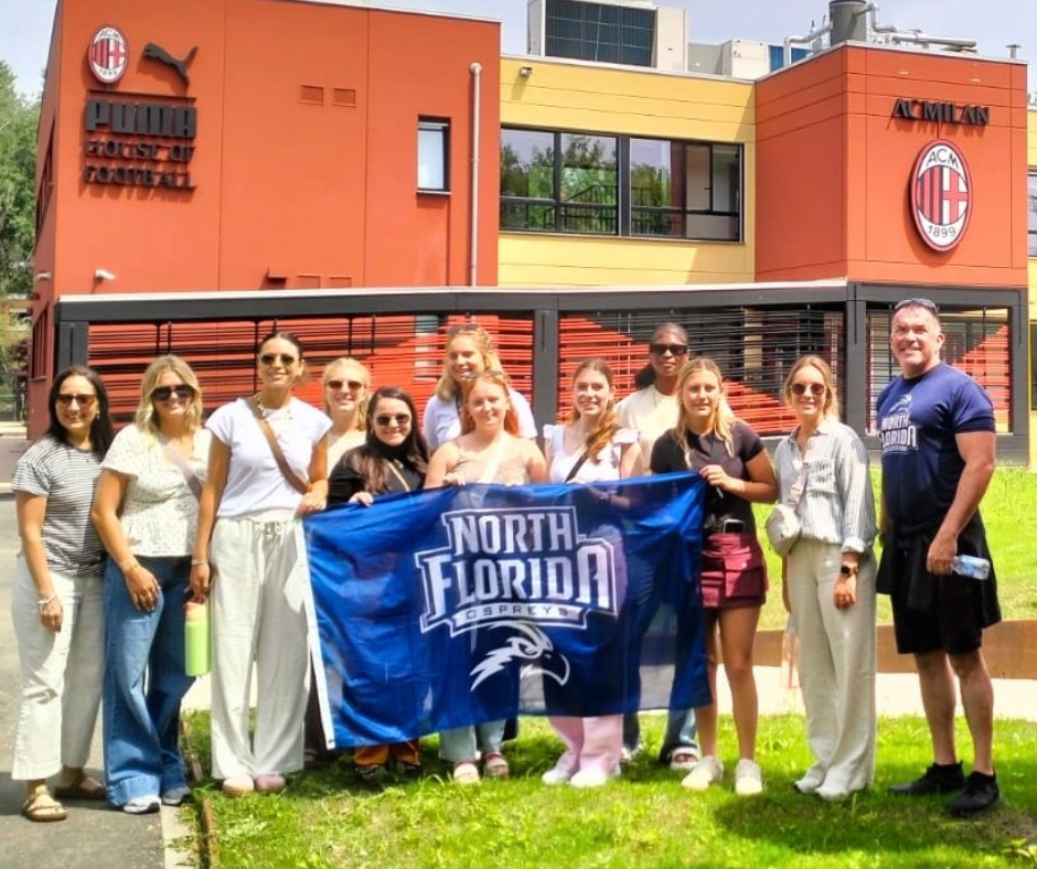 A group of students and faculty stand holding a UNF banner in Italy.