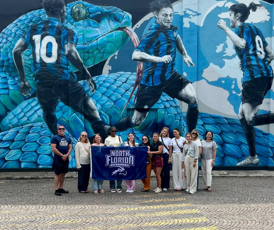 A group of students and faculty stand holding a UNF banner in Italy.