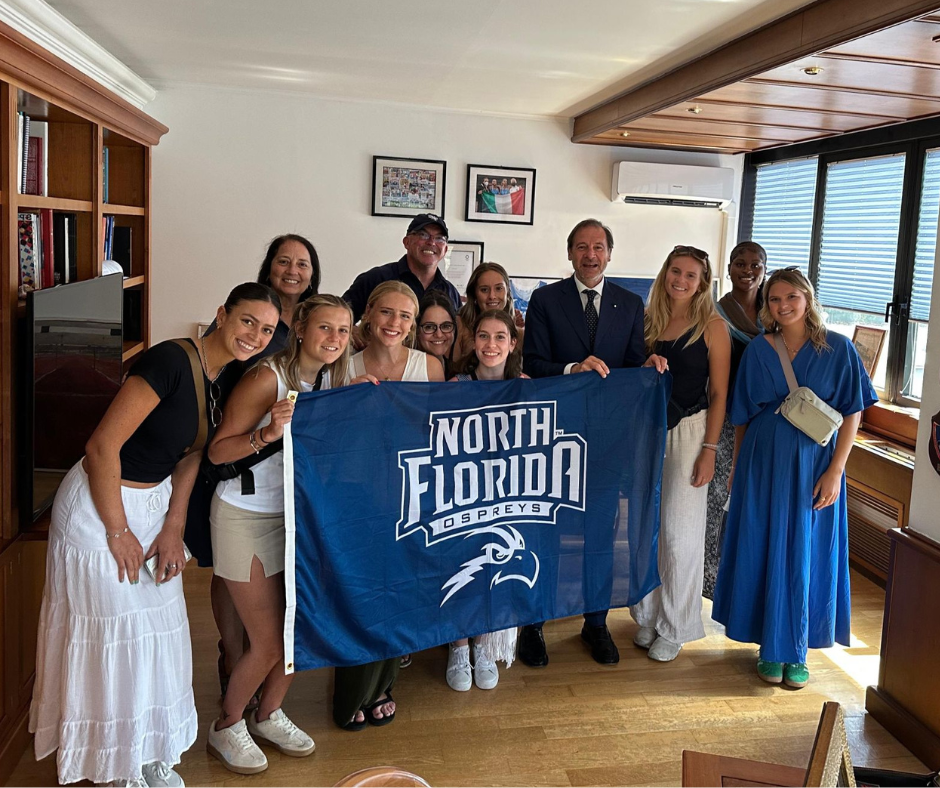 A group of students and faculty stand holding a UNF banner in Italy.
