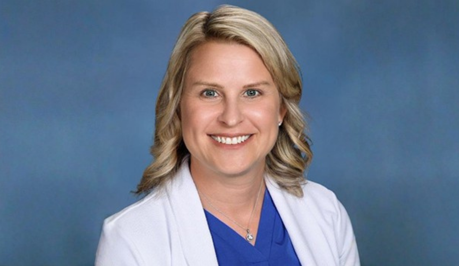 Professional headshot of a smiling woman with blonde hair wearing a white coat over blue scrubs against a blue background.